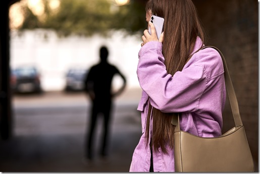 Criminal stalking woman, commiting crime while victim was walking alone, talking on phone in dark street. Caucasian young woman is looking back, afraid of male stranger person in the background