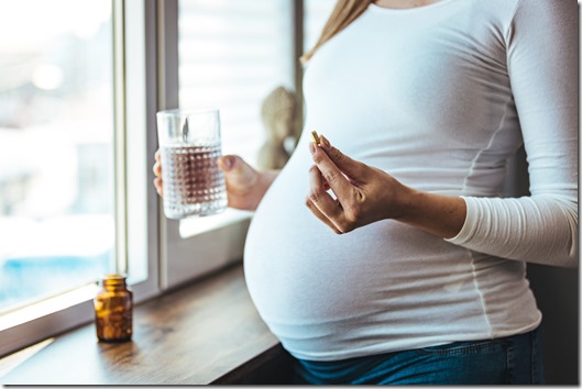 Pregnant woman reading label on bottle with medicine, with vitamins. Female sitting at home in bed with glass of water medicine. Pregnancy, health, pharmaceuticals, care and people.