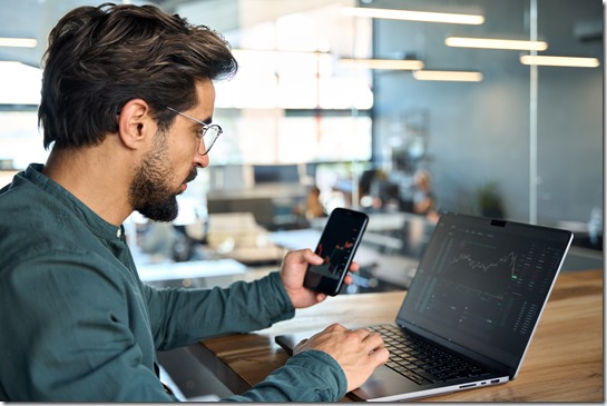 Stock trader investor holding phone using laptop analyzing trade market data.